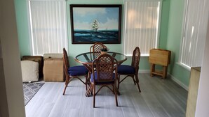 Een eetruimte met een ronde glazen tafel, vier houten stoelen met blauwe kussens, en een schilderij van een schip op zee.