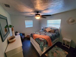 Une chambre à coucher comprenant un lit, un ventilateur de plafond, deux fenêtres avec stores, une table de chevet avec une lampe, un petit tabouret et un chapeau décoratif accroché au mur.