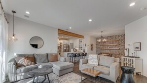 Ein modernes Wohnzimmer mit einem grauen Ecksofa, einem weißen Sessel und einem Couchtisch. Im Hintergrund ist eine Küche erkennbar, mit einer Ziegelwand und Pendelleuchten.