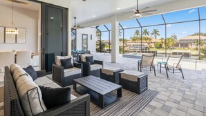 Un vaste espace extérieur aménagé avec des meubles modernes, de grandes fenêtres offrant une vue sur les palmiers et une piscine, ainsi que des ventilateurs de plafond assurant la ventilation.