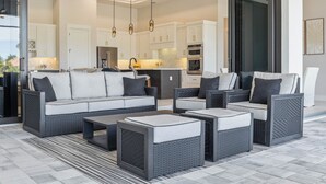 Un espace de vie moderne comprenant un ensemble de canapés, une table basse et des fauteuils, une cuisine avec des meubles blancs et des appareils électroménagers, et une grande porte vitrée donnant sur un espace extérieur.