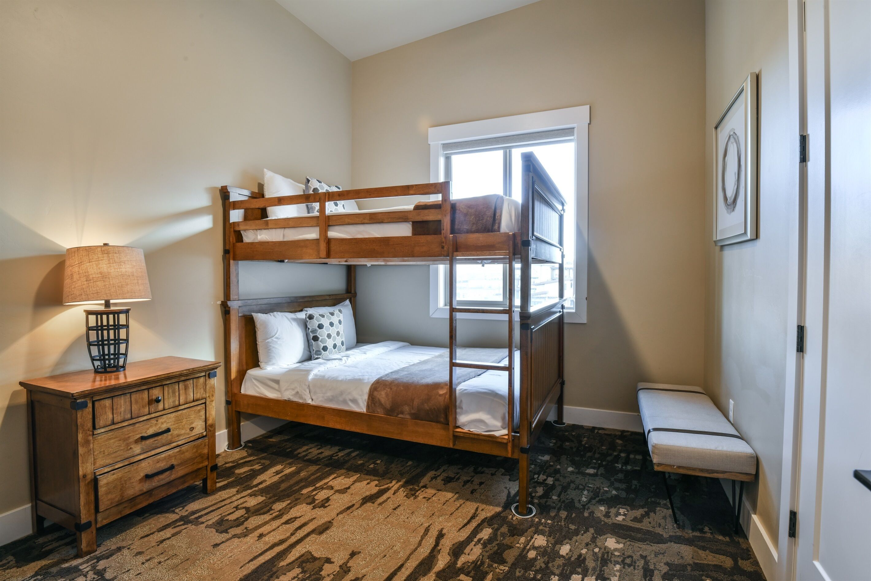 A bunk bed room with a wooden dresser, a small bench, and a window providing natural light.