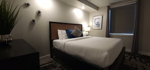 Een hotelkamer met een groot bed, een nachtkastje, een lamp, decoratieve wandkunst en een raam met gordijnen.