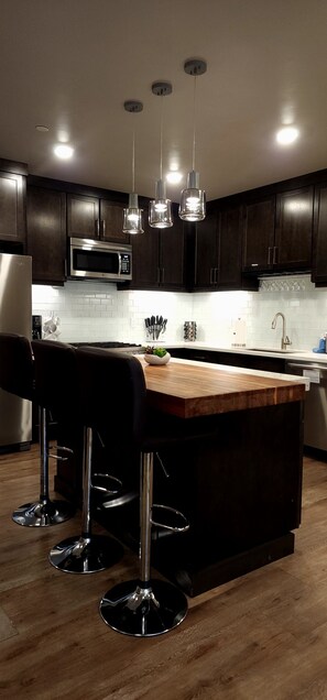 A modern kitchen with dark wooden cabinets, stainless steel appliances, a wooden island with bar stools, and pendant lighting. - D1 and D4 Combo Unit - Sage Creek Resort Moab (Moab)