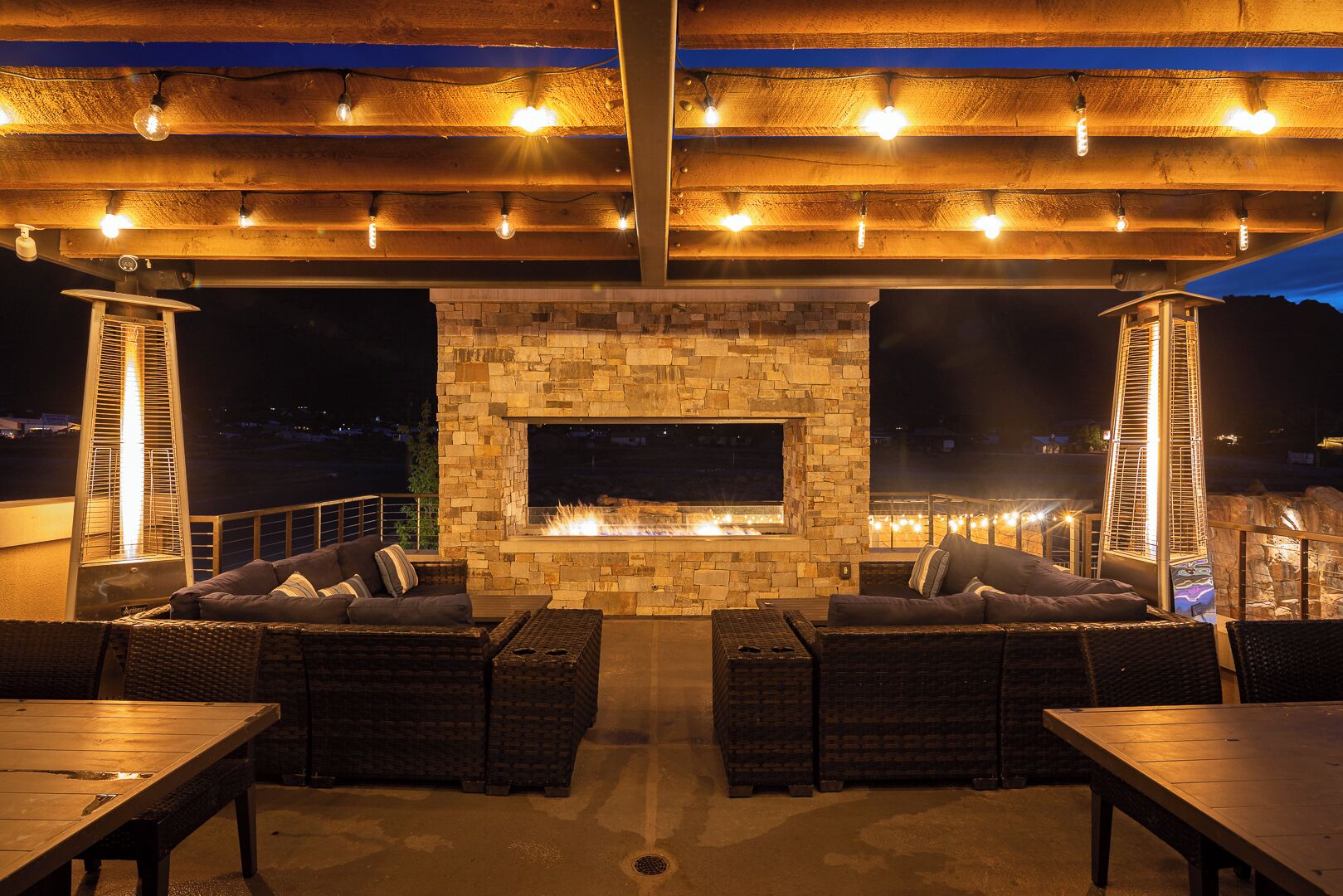 Une terrasse extérieure avec des murs en pierre, une cheminée, des meubles en osier et des guirlandes lumineuses.