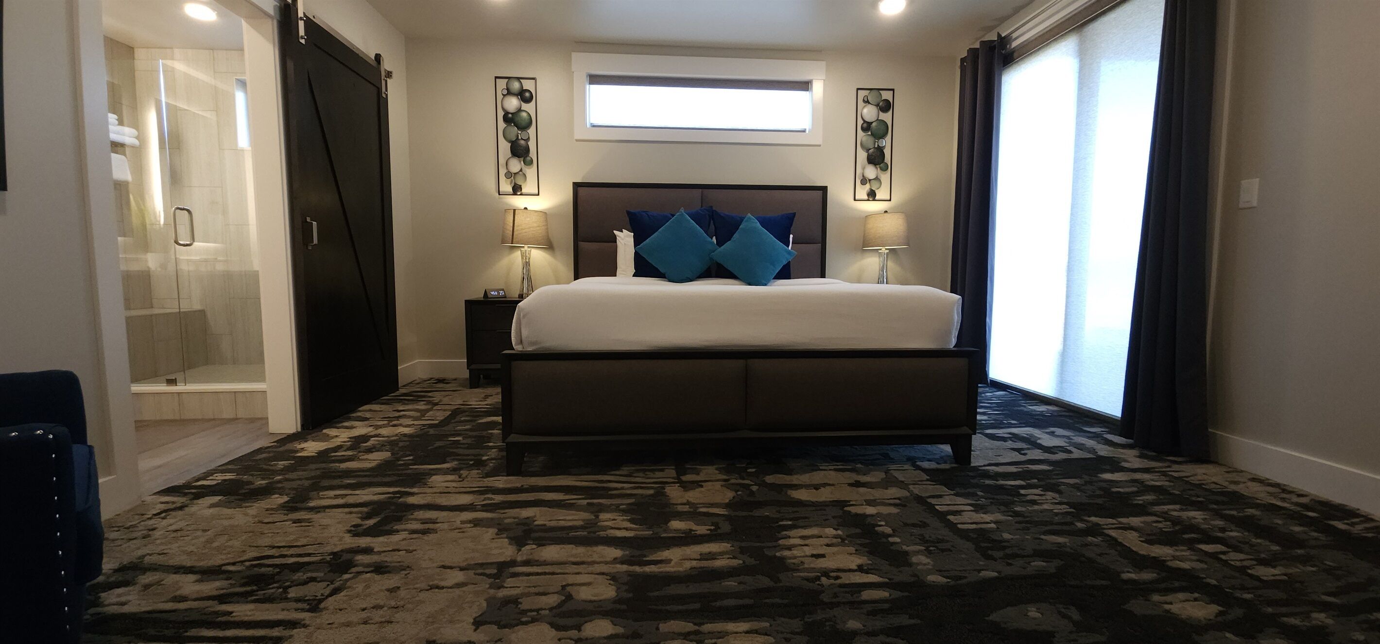 Une chambre d’hôtel avec un grand lit, deux tables de chevet avec des lampes, une tête de lit sombre, une moquette à motifs, une fenêtre avec des rideaux et une œuvre d’art accrochée au mur.