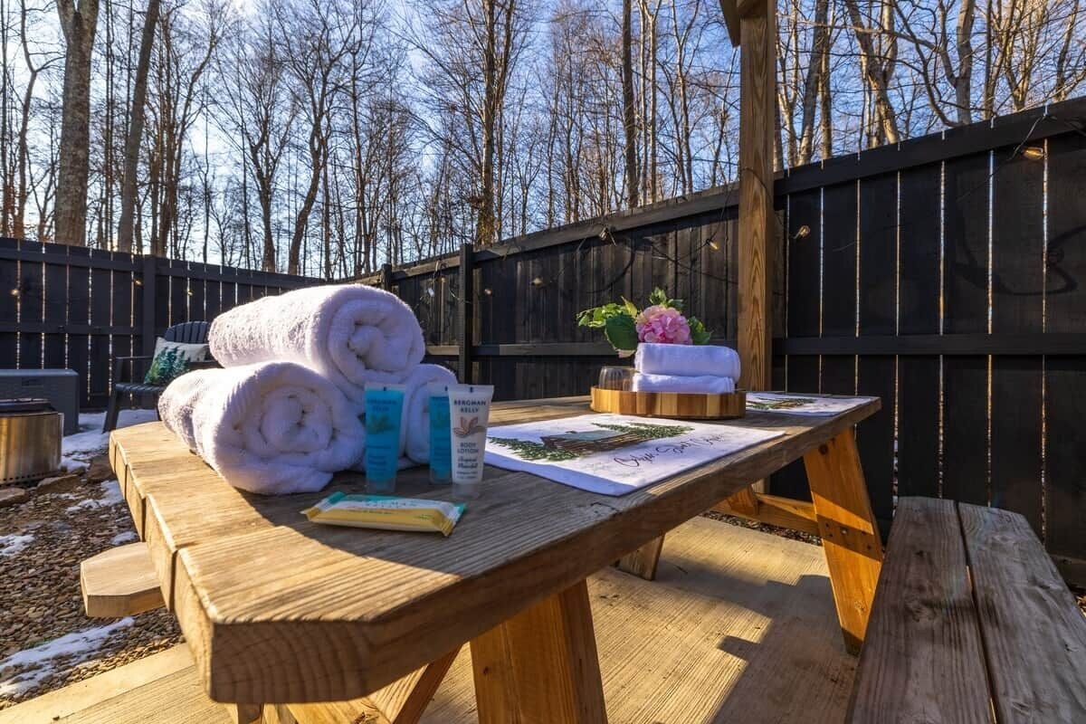 A wooden picnic table with towels, a bottle, and a card in a backyard setting.