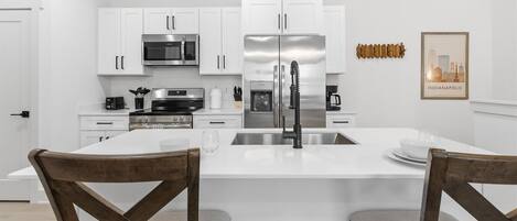 Une cuisine moderne avec des meubles blancs, des appareils en acier inoxydable et un îlot central agrémenté de deux chaises en bois.