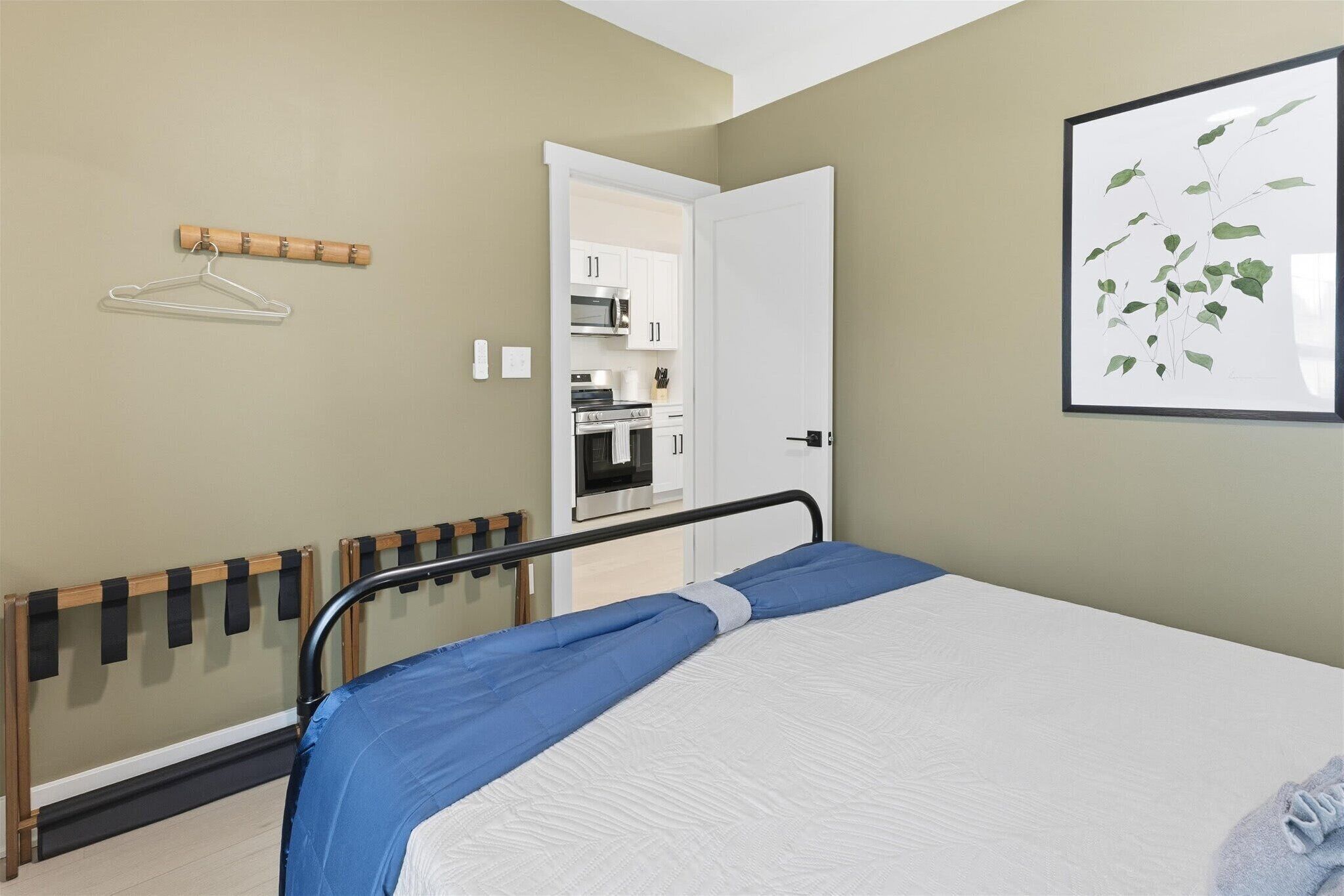 A bedroom with a bed, a hanging clothes rack, a door to a kitchen, and a framed picture on the wall.