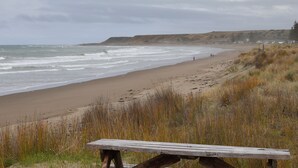 Ein Strand mit einer Holzbank im Vordergrund, Wellen, die auf die Küste treffen, und eine hügelige Landschaft im Hintergrund.