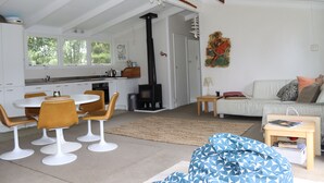 Eine moderne Küche mit weißen Schränken, ein Essbereich mit rundem Tisch und Stühlen, ein Wohnzimmer mit Sofa und einem blauen Sitzsack mit Muster, und ein Kamin.