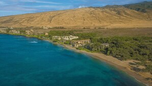 Vue aérienne d’une zone côtière avec une plage, un grand ensemble de bâtiments et un paysage vallonné en arrière-plan.