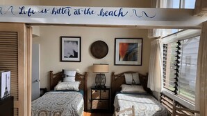 Une chambre confortable avec deux lits, une petite table et une chaise. Les murs sont ornés de tableaux encadrés, il y a une lampe et une fenêtre avec des stores.