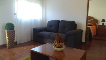 Una sala de estar con un sofá oscuro, una mesa de centro de madera, un tapete verde, una maceta de mimbre con un árbol pequeño y una ventana con cortinas de encaje.