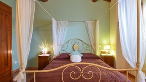 Uma cama de dossel com estrutura de madeira e cabeceira com padrão floral. Há duas mesas de cabeceira com luminárias e um criado-mudo com abajur. O quarto possui teto de madeira e paredes com tom esverdeado.