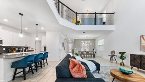 A modern kitchen with white cabinets, a bar area with blue chairs, a dark sofa with a throw blanket, and a wooden coffee table. - 5BR 5BA Home Storey Lake Water Park Amenities (Kissimmee)