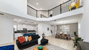 A spacious living area with a black sectional sofa, a wooden coffee table, and a dining table with chairs. A kitchen is visible in the background, and there is a staircase leading to an upper level. - 5BR 5BA Home Storey Lake Water Park Amenities (Kissimmee)