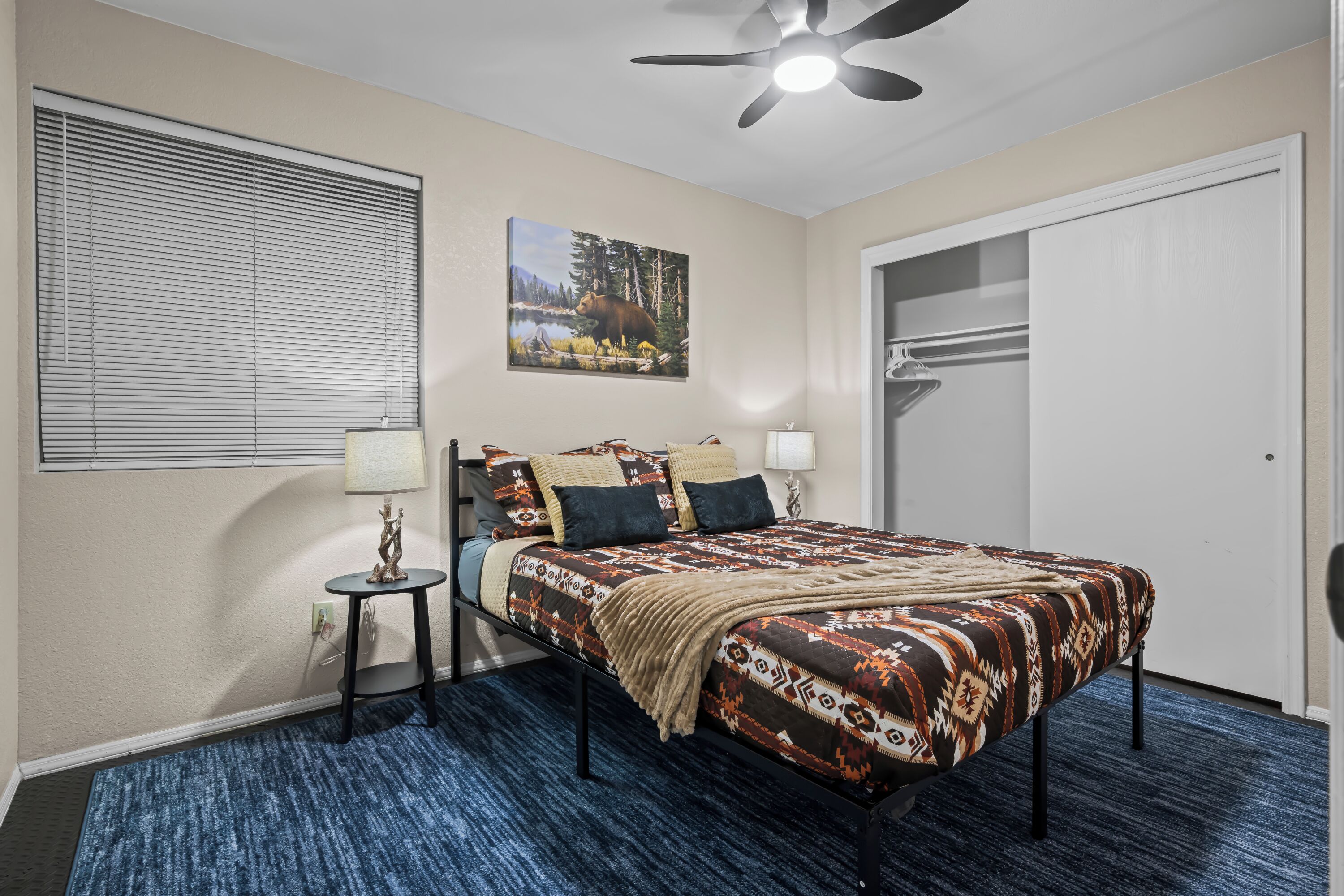 A bedroom with a bed, a ceiling fan, a window with blinds, a painting, a small table with a lamp, and a closet.