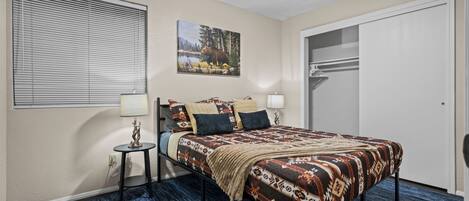 A bedroom with a bed, a ceiling fan, a window with blinds, a painting, a small table with a lamp, and a closet.