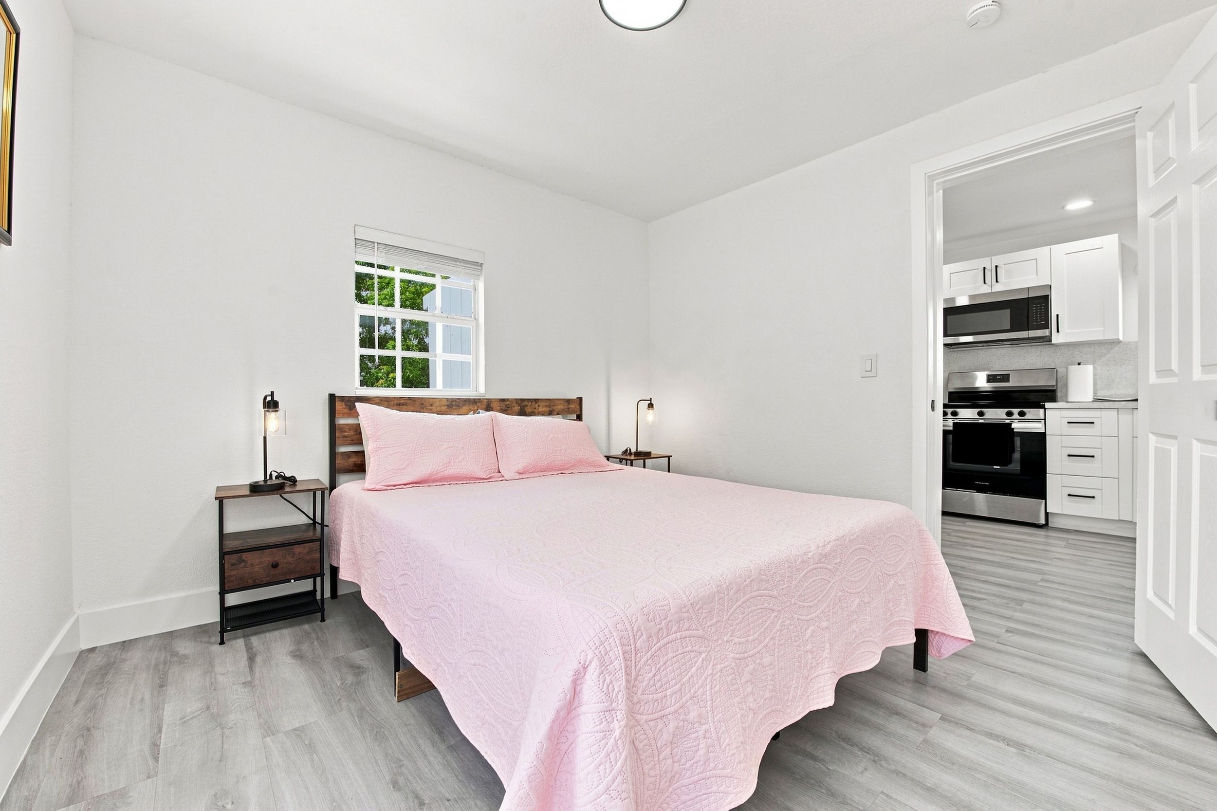 A bedroom with a bed, bedside tables, a window, and a view of a kitchen.
