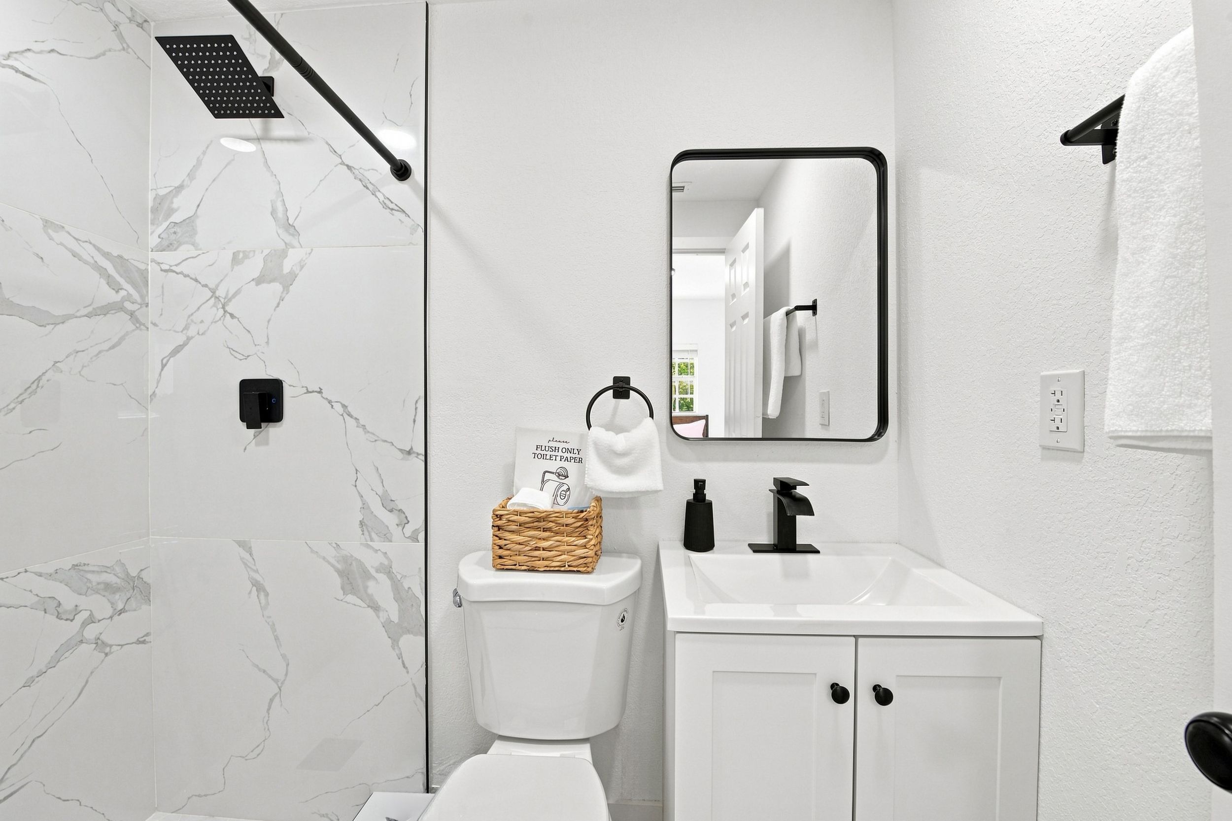 Un baño moderno con pared de mármol blanco, un inodoro, un lavabo con grifo negro, un espejo y zona de ducha.