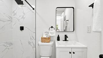 Un baño moderno con pared de mármol blanco, un inodoro, un lavabo con grifo negro, un espejo y zona de ducha.