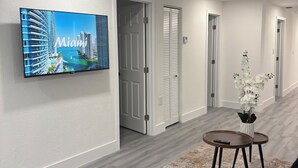 A modern hotel lobby with a flat-screen TV displaying a cityscape, a black sofa, a small round table with a vase of flowers, and a patterned rug. - La Orquidia (Miami)