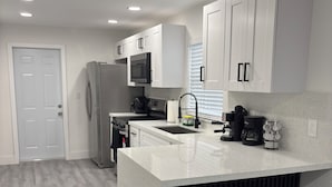 A modern kitchen with white cabinetry, a stainless steel refrigerator, a microwave, and a coffee machine. There are two stools under the island and a window with blinds. - La Orquidia (Miami)