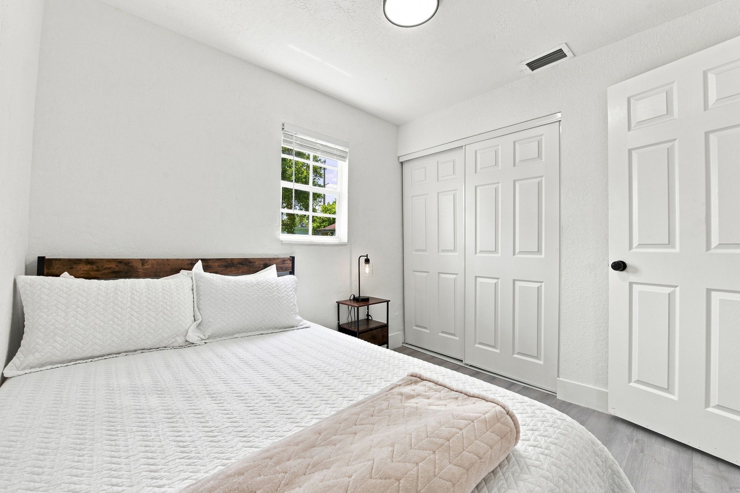 Un dormitorio con una cama, una mesita de noche, un ventanal y un conjunto de puertas blancas.