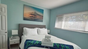 A neatly made bed with white linens, a blue and green throw blanket, a nightstand with a lamp, a painting of a mountain landscape, and a window with blinds. - Charming Bright 3BR House - Upper Level Suite (Burnaby)