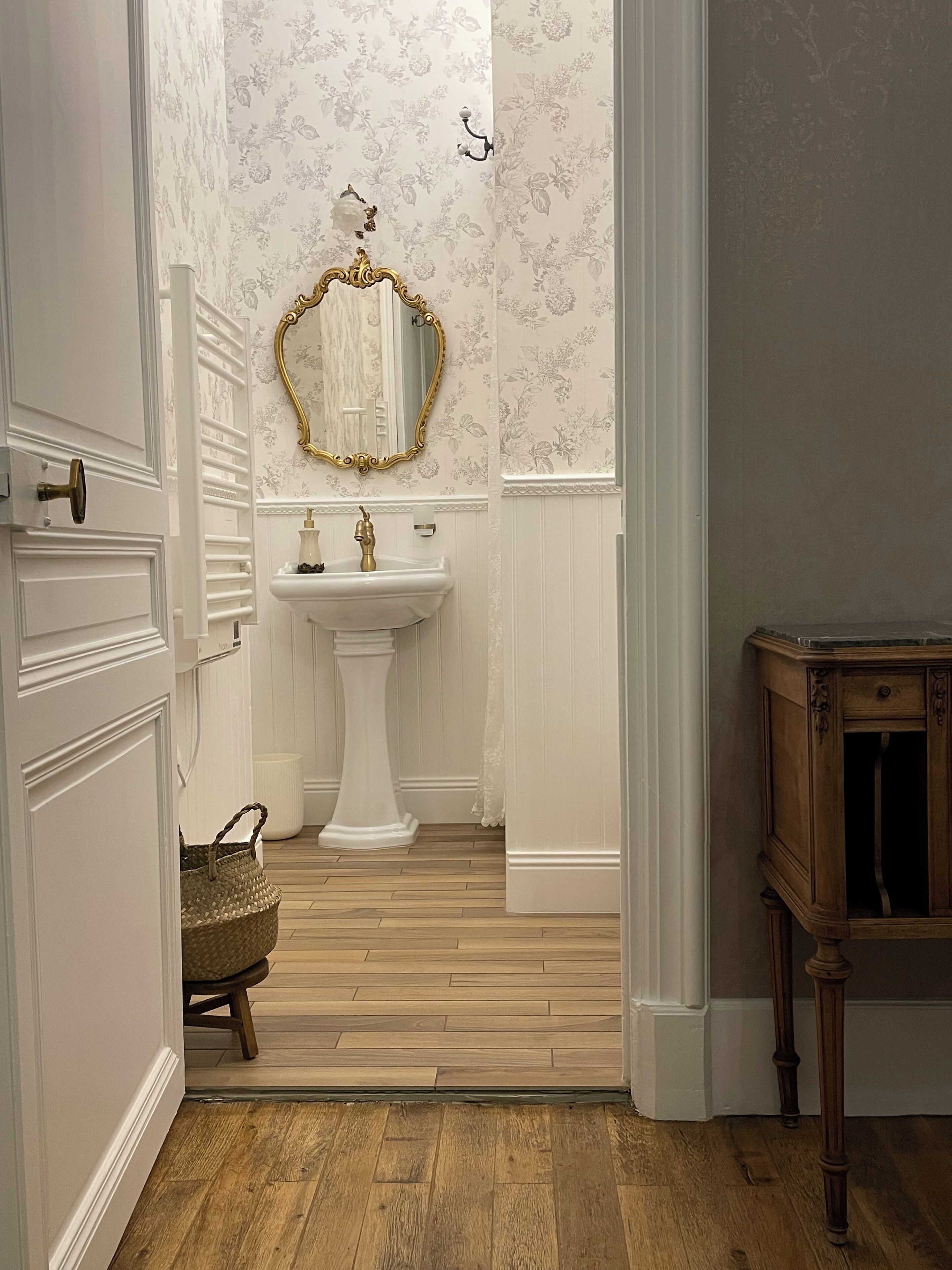 A hallway leading to a bathroom with a white pedestal sink, a gold-framed mirror, and floral wallpaper.