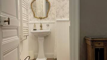 A hallway leading to a bathroom with a white pedestal sink, a gold-framed mirror, and floral wallpaper.