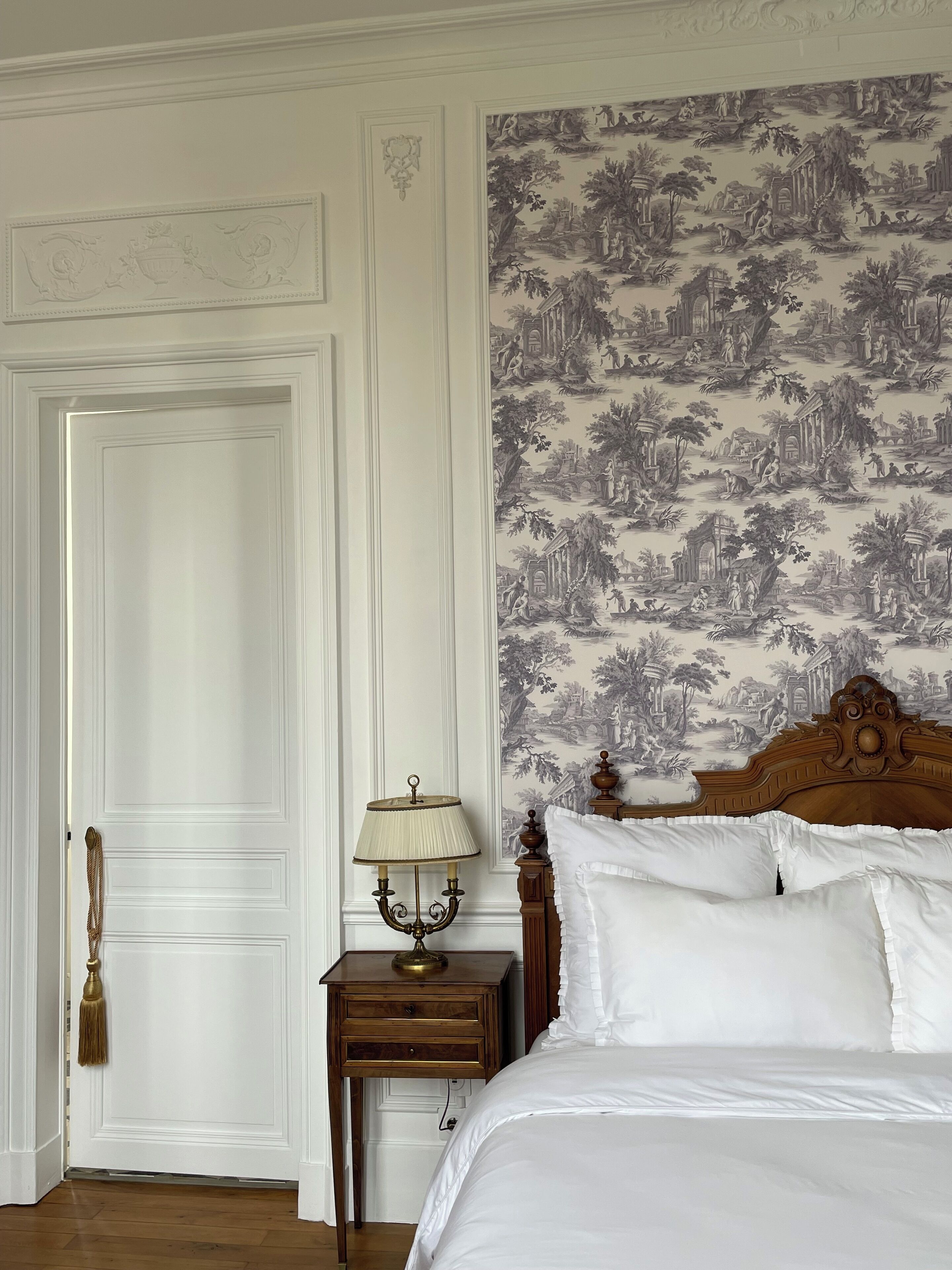 Un dormitorio con una puerta blanca, una mesita de noche con lámpara, una cama con ropa de cama blanca y un papel tapiz con motivos naturales.