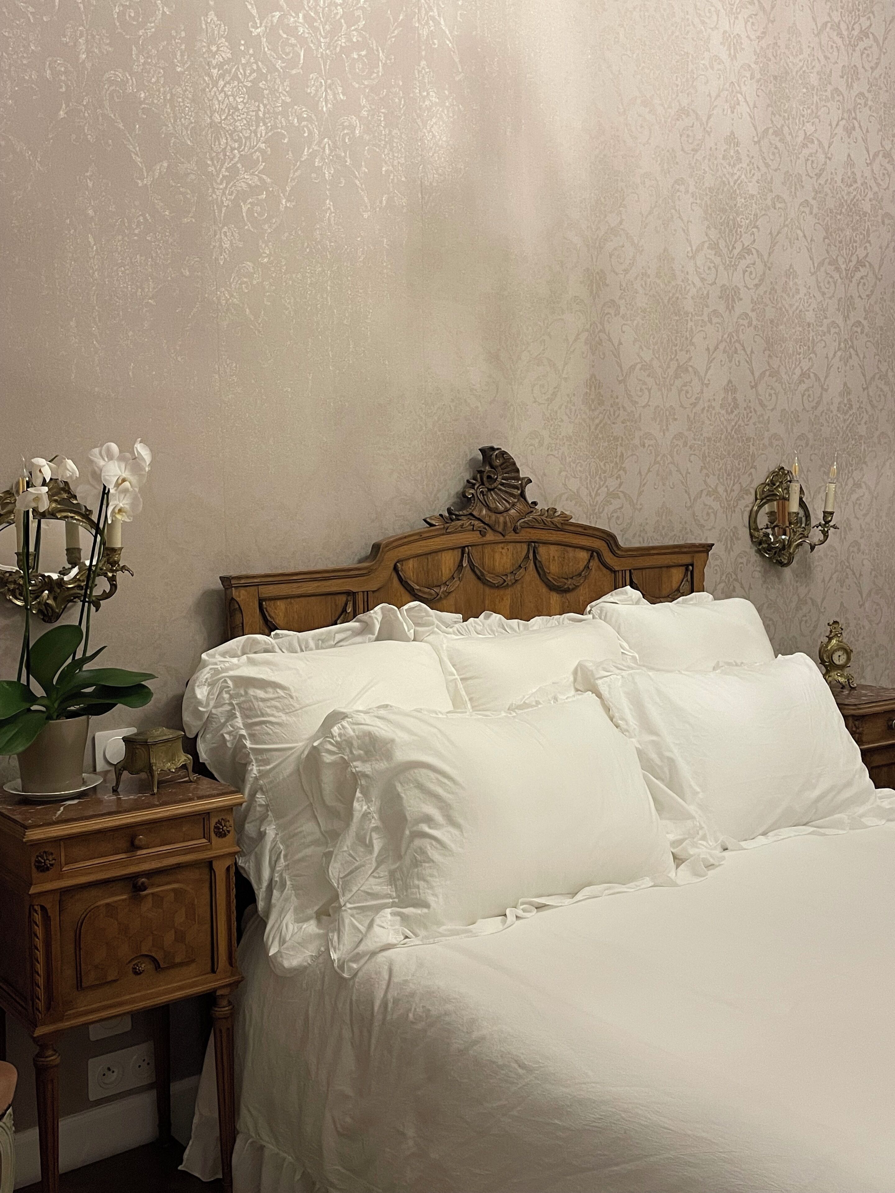 A neatly made bed with white linens, a wooden headboard, a bedside table with a plant, and a wall-mounted light fixture.