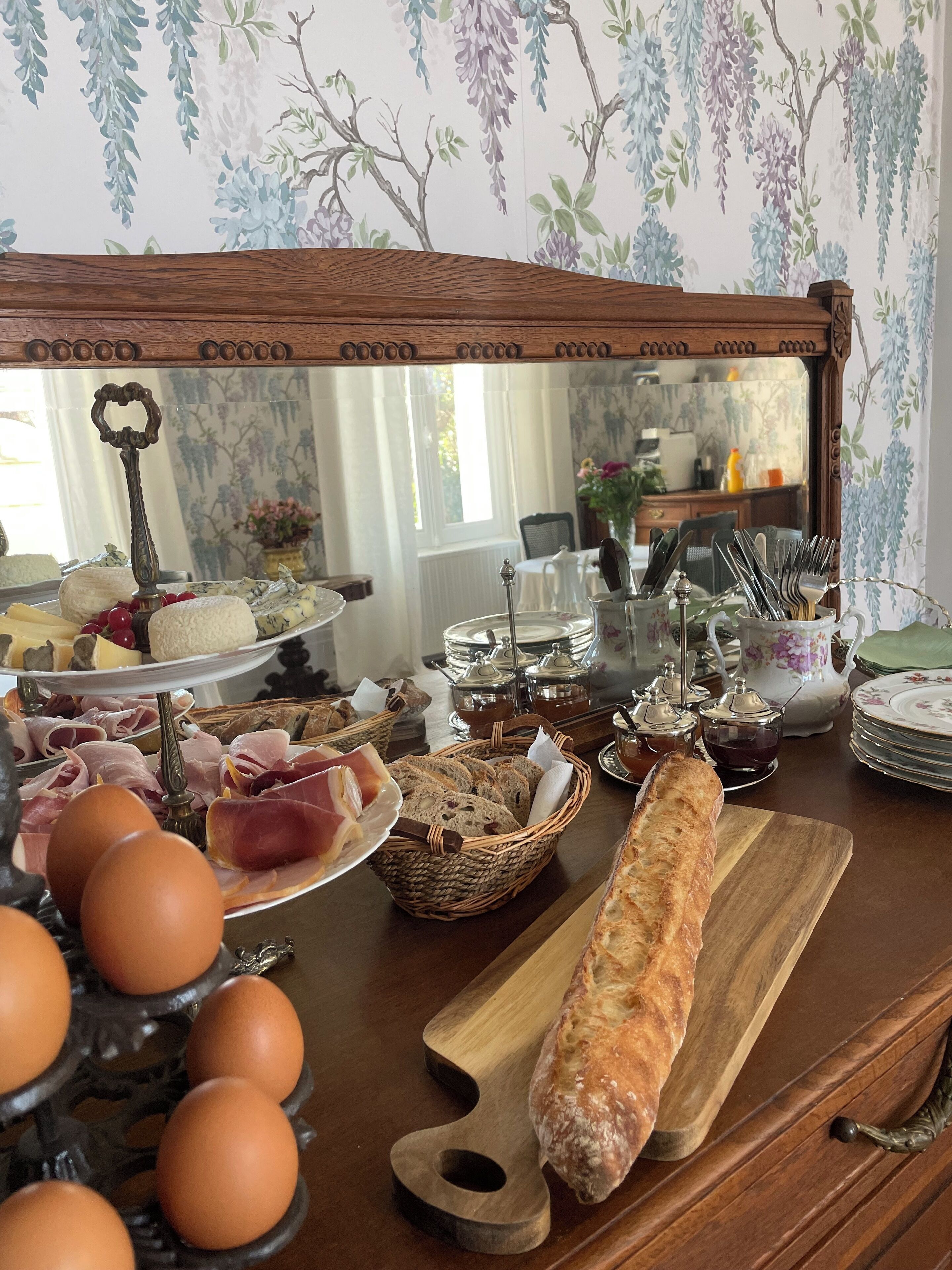 A breakfast buffet with various food items, including eggs, bread, and cheese, arranged on a wooden table with a floral wallpaper background.