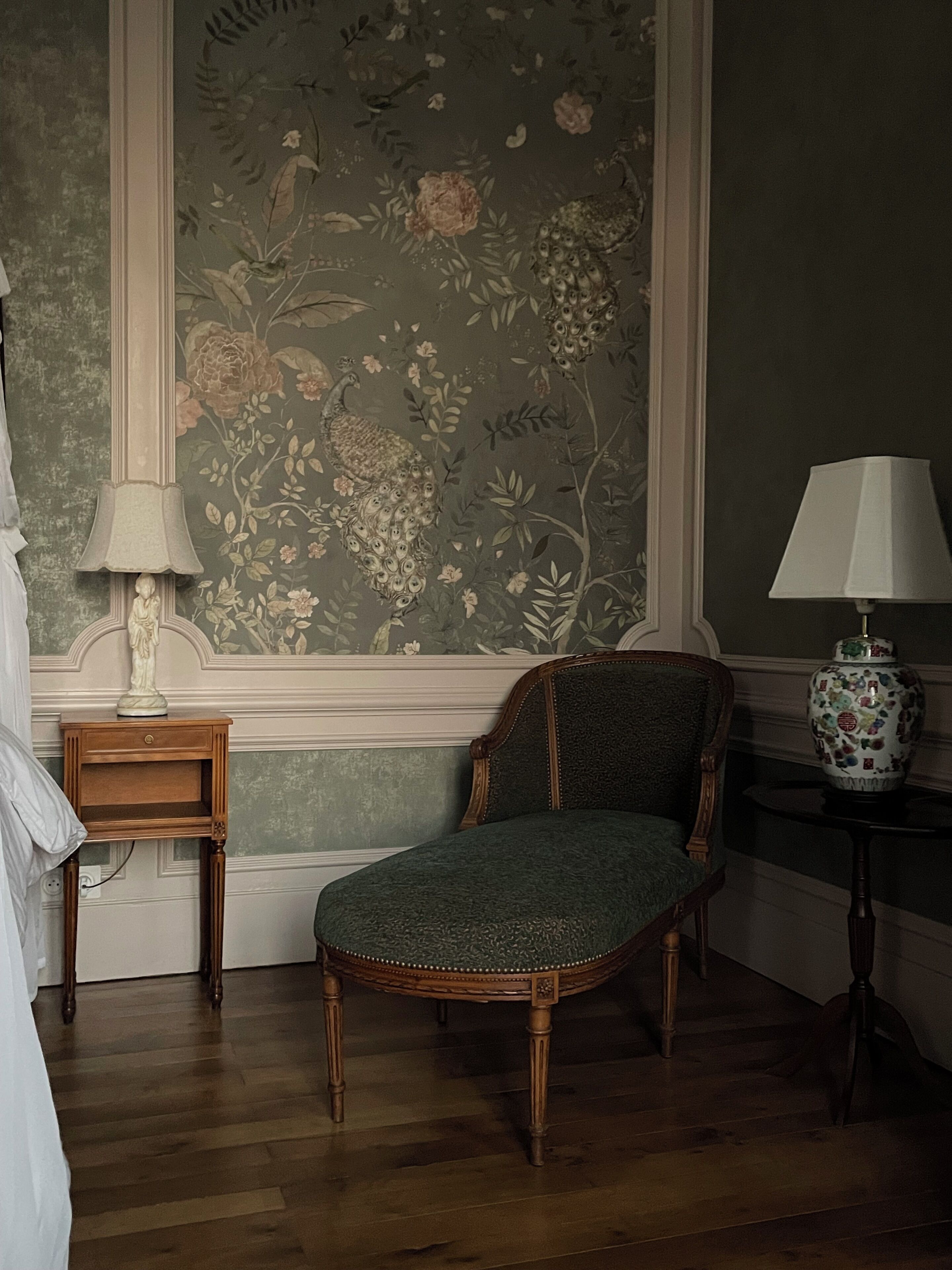 Una habitación con una silla tapizada en verde, una mesita de noche de madera con una lámpara, y un papel pintado con motivos florales.