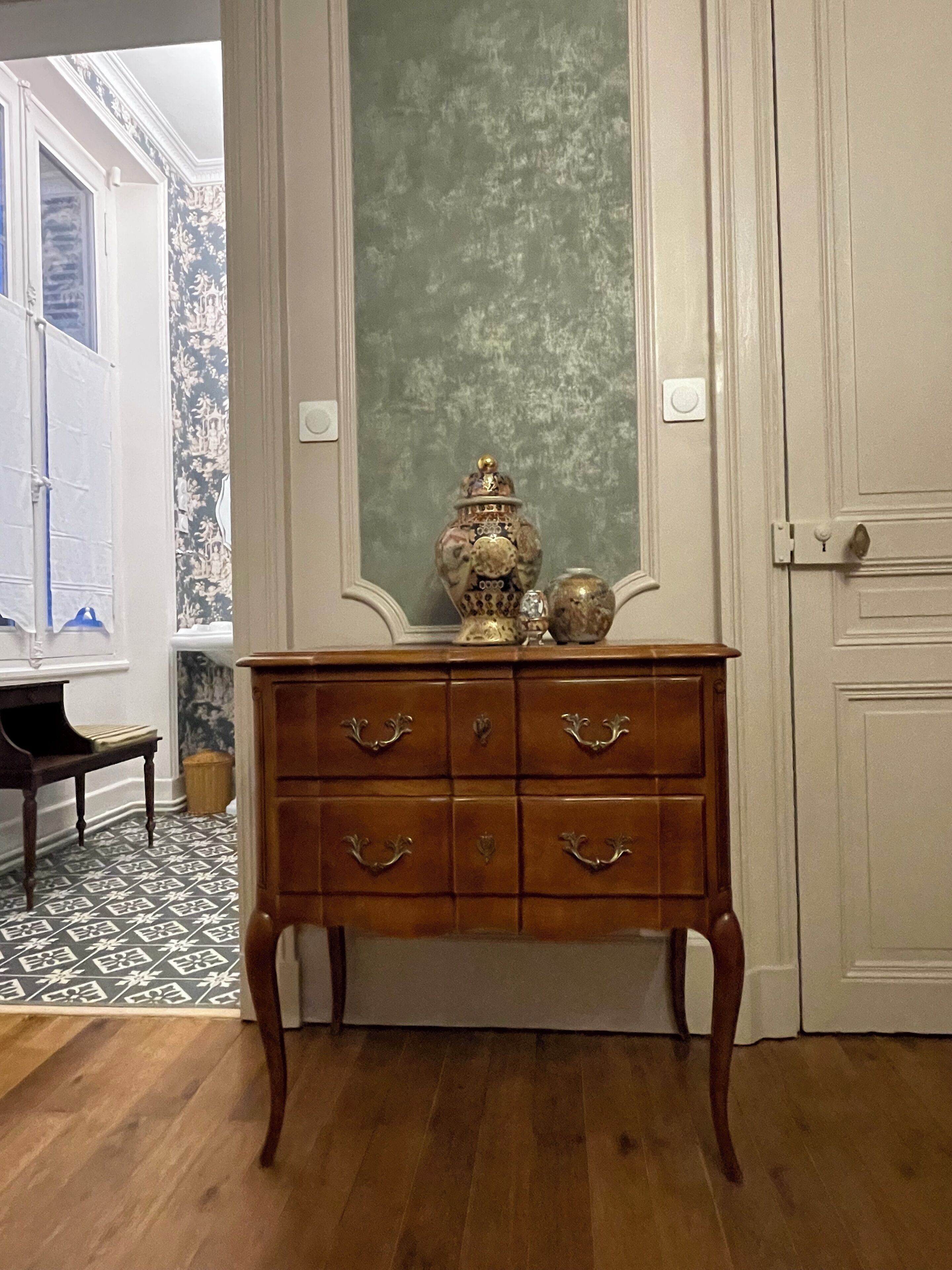 A wooden dresser with six drawers, a mirror, and a decorative vase in a room with patterned wallpaper and a wooden floor.