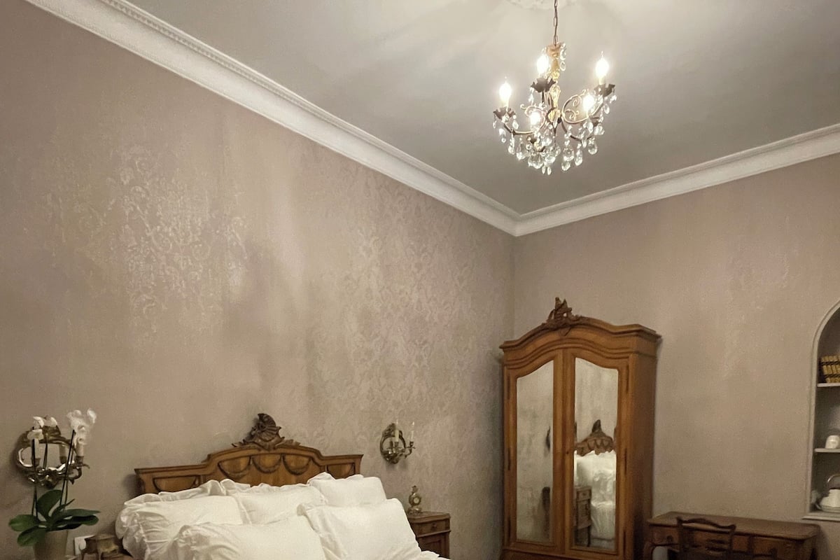 Un dormitorio con una cama grande, suelo de madera, una araña, un armario de madera y un escritorio con silla.
