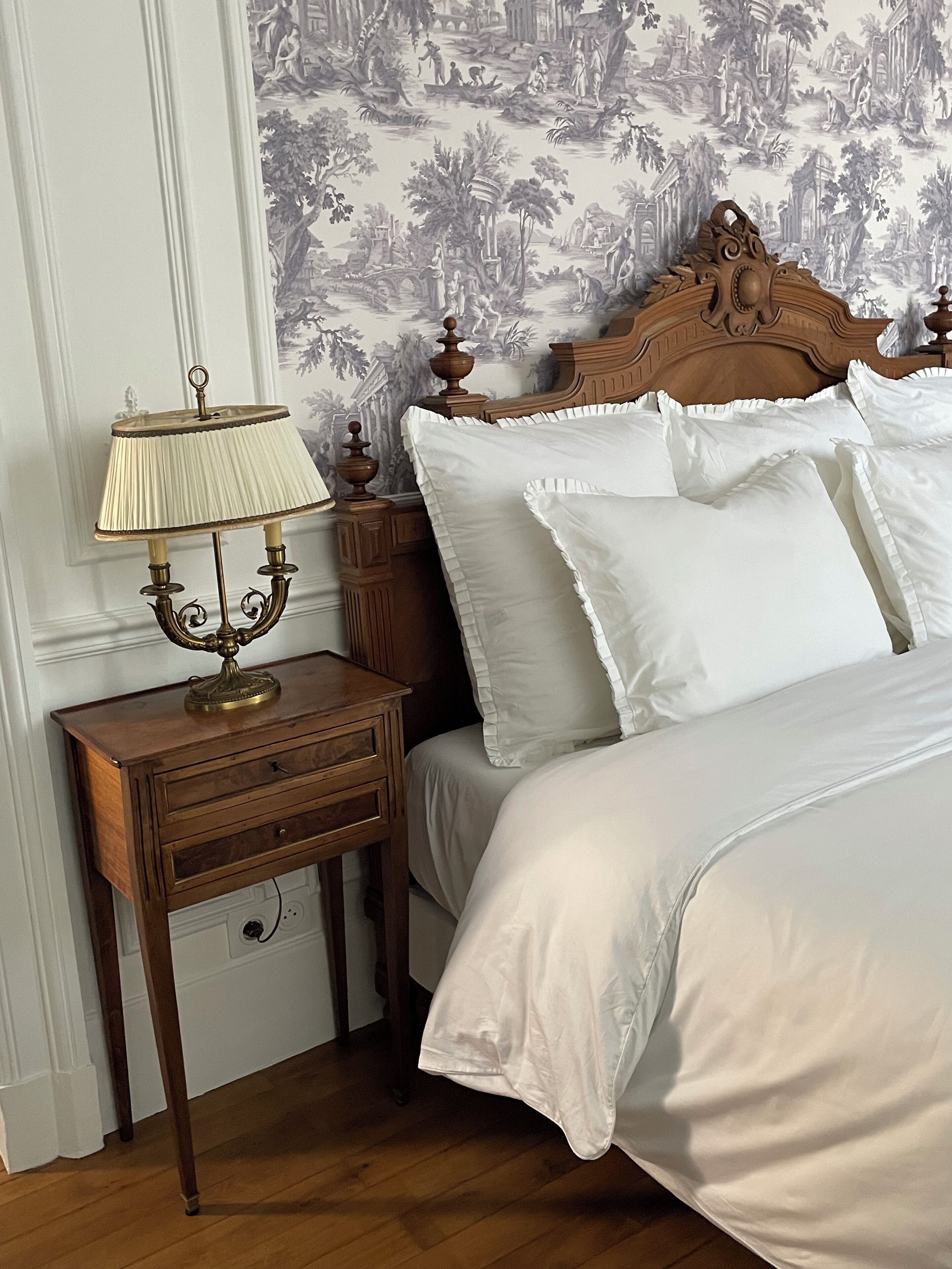 Een netjes opgemaakt bed met witte lakens en een houten hoofdbord, een nachtkastje met een lamp, en een wand met behang met een natuurmotief.