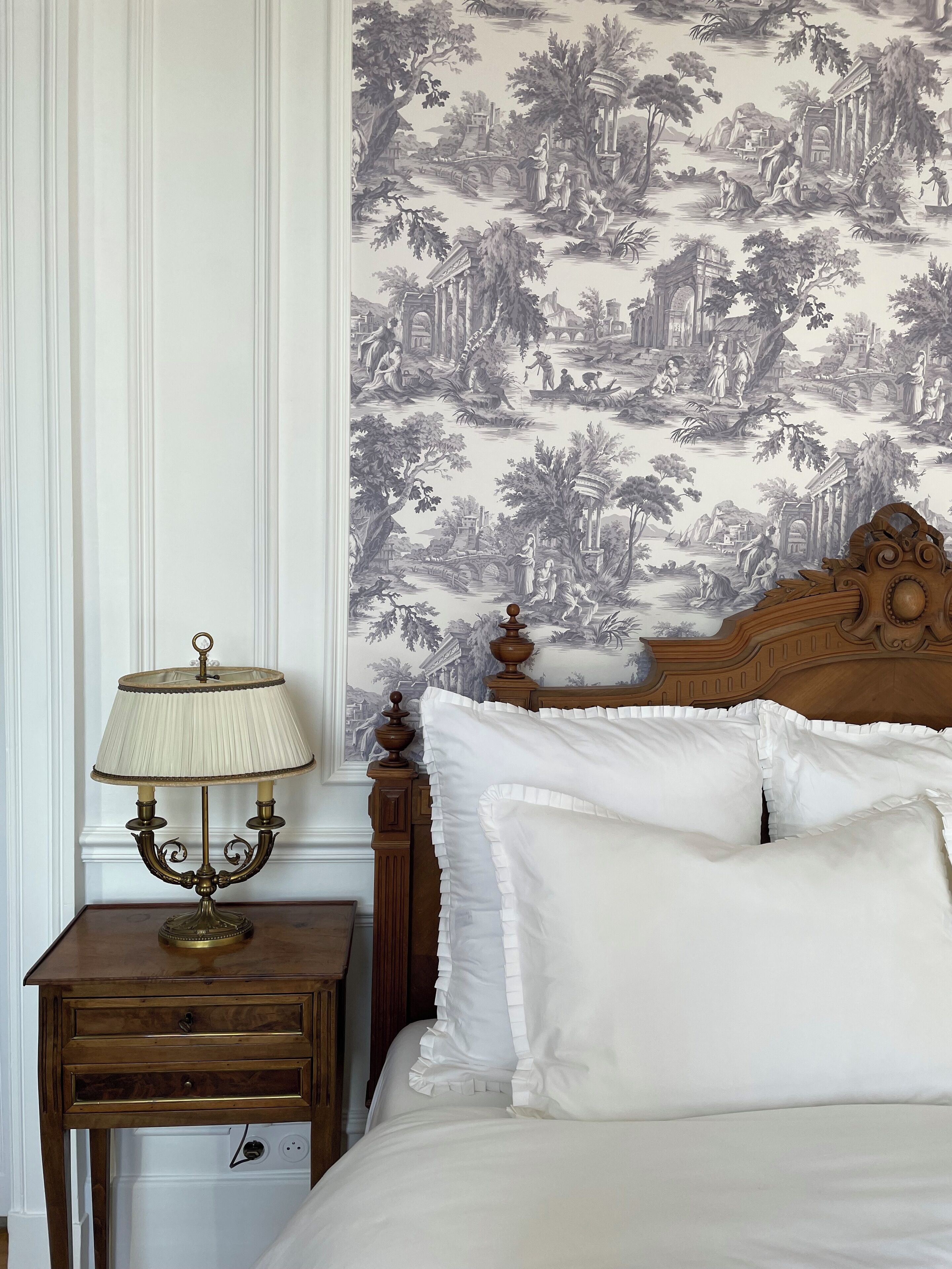 A bedroom with a wooden headboard, a bedside table with a lamp, and a wall with a nature-themed wallpaper.