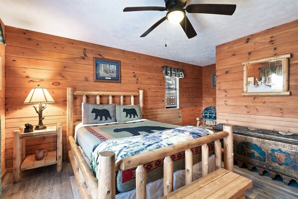 Un dormitorio de cabaña de madera con una cama, una mesita de noche, una lámpara, un ventilador de techo y un espejo decorativo.