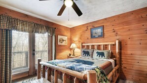 Une chambre dans une cabane en bois, avec un lit, un banc, un ventilateur de plafond et une fenêtre avec des rideaux.