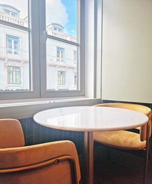 A corner of a room with a round table and two chairs, a window with a view of buildings, and a tiled floor.