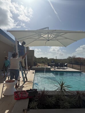 Ein Schwimmbecken mit einer Terrasse, einem weißen Sonnenschirm und einigen Personen in der Nähe.