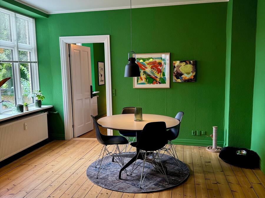 Una habitación con una mesa redonda, sillas negras, una lámpara colgante negra, un cuadro abstracto enmarcado, una ventana con persianas y un radiador.