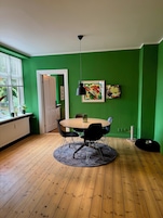 Una habitación con una mesa redonda, sillas negras, una lámpara colgante negra, un cuadro abstracto enmarcado, una ventana con persianas y un radiador.