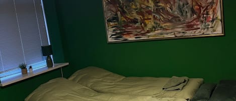 Una habitación con cama, pared verde, un cuadro y ventana con persianas.