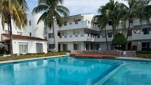 A swimming pool with a stone border, surrounded by white buildings with balconies and palm trees. - Casas Arrecife (Ixtapa)
