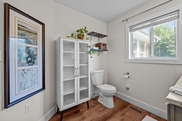 A bathroom with a toilet, a cabinet with glass doors, a window, and a framed picture on the wall. - Secluded Horse-Country Farmhouse w/ Pond+36 Acres+Near Durham (Rougemont)
