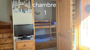 Een kamer met stapelbedden, een houten kast met een televisie, een wandklok en een deur met de tekst 'chambre 1'.